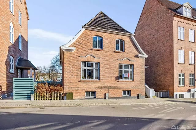 A red brick house stands quietly between two larger buildings. The house has a sloping roof and several windows. It is located on an urban street with sidewalks and a road in the foreground.