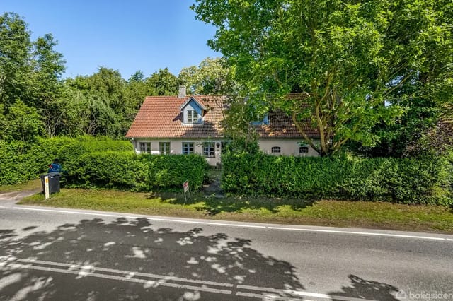 House with red tile roof behind green hedge, surrounded by trees and bushes
