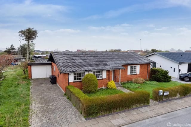 A red brick house with a black roof along a paved driveway, surrounded by a well-kept lawn and hedges under a clear blue sky in a suburban area.