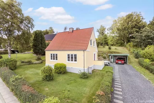 A yellow house surrounded by a garden with bushes and trees. To the right is a gravel road with a carport where a red car is parked.