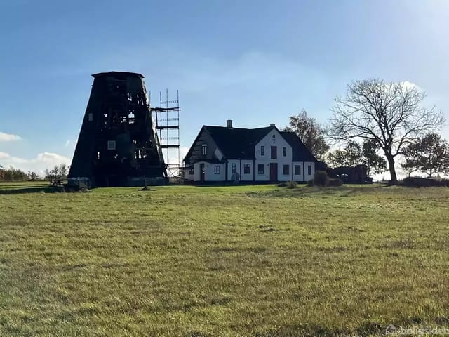 An old windmill under renovation next to a traditional white house in an open, green landscape with scattered trees and a clear blue sky.