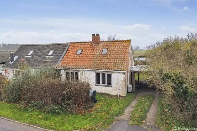 House with a gabled roof in disrepair, next to another house, surrounded by bushes, on a lawn, with a driveway and road in the foreground.