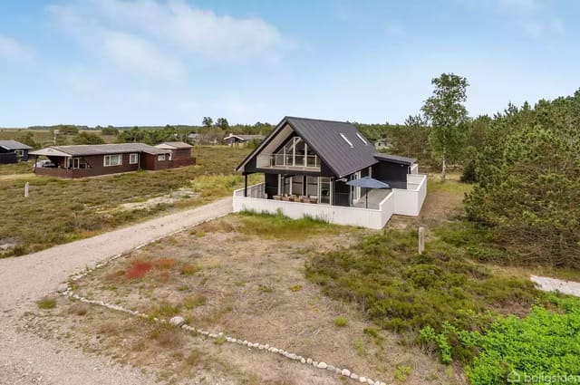 A modern house with a black roof and white walls stands on an open plot. It is surrounded by low bushes and gravel paths in a rural area.