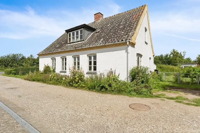 White brick house with black roof, situated on a gravel road surrounded by grass and small bushes. Blue sky and green trees in the background.