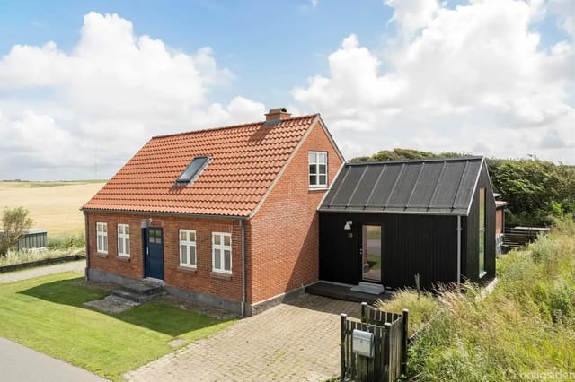 A brick house with a red tile roof stands next to a modern black annex on a well-kept lawn with a driveway. Fields and a blue, cloudy sky are in the background.