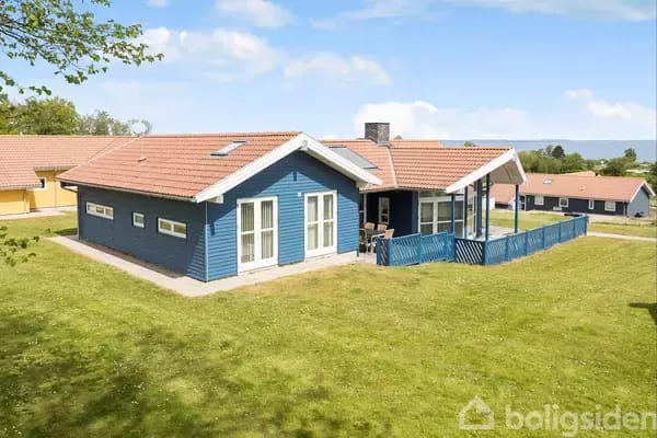 Blue house in a green garden surrounded by grass and other houses in the background. The house has white doors and windows and a terrace with table and chairs.