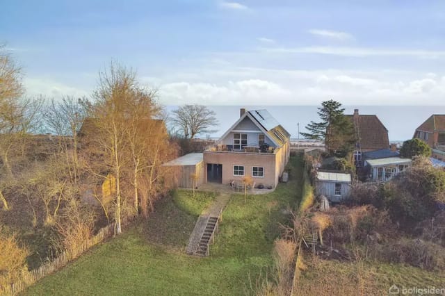 A house with a sloped roof and solar panels stands in a lush garden with a staircase. In the background, several houses and a sea view are visible.