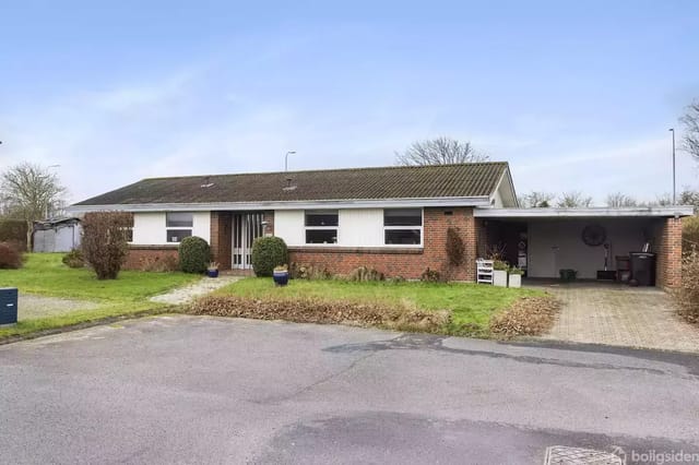 A single-story brick house with a flat roof stands quietly on a calm residential street. In front of the house is a well-maintained lawn and a driveway with a carport to the right.