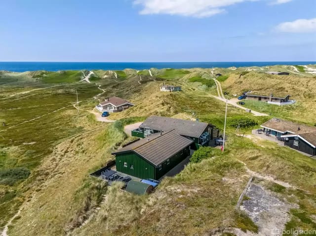 House surrounded by green dunes, with a coastline and blue sea in the background.