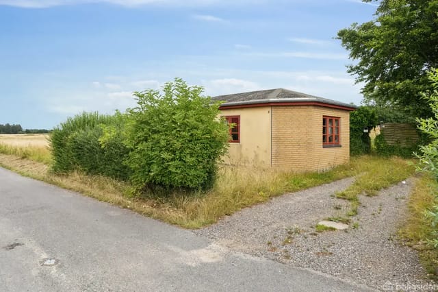 A low, square building with red windows stands quietly surrounded by tall grass and bushes at a country road intersection. Blue sky and open fields in the background.