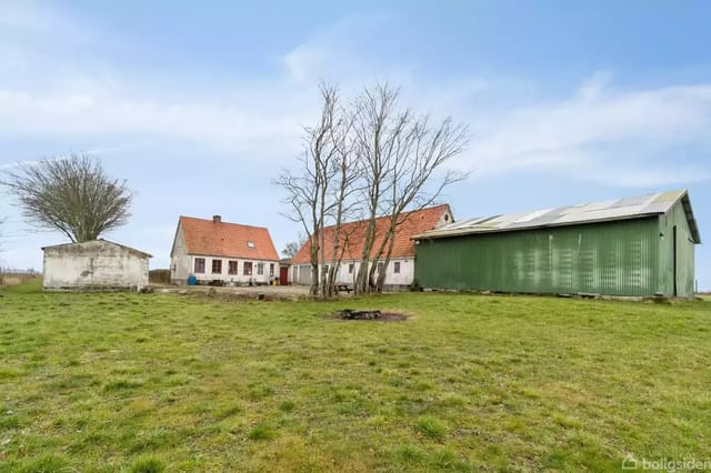 A farm with a red tiled roof and a green barn surrounded by a lawn. Trees are placed in the middle of the image under a blue sky.