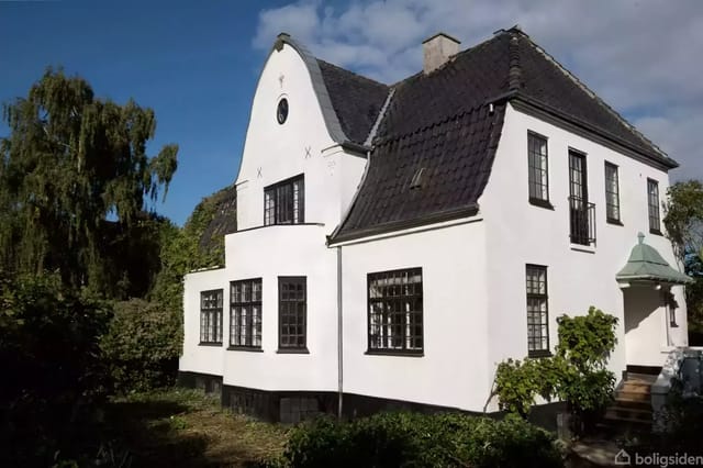 White two-story house with black slate roof stands still in a lush garden. Large windows frame the facade, and a small staircase leads up to the front door.