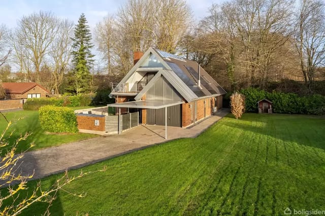 Two-story villa with large glass sections and sloping roof, surrounded by a well-maintained lawn and trees, located in an open, green area. A carport is connected to the main building.