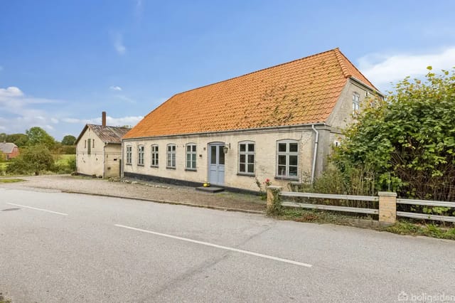 House with red tile roof and white walls by a country road, surrounded by green bushes and lawns under a clear blue sky.