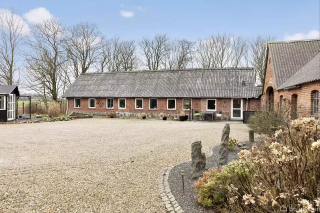 A long, low brick building with a gray roof stands centrally in the image. The building is surrounded by a gravel courtyard and rustic gardens. The background has leafless trees.