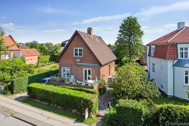 A brick house with a sloped roof stands in a well-kept garden surrounded by green bushes. In front of the house is a wooden terrace with garden furniture. It is located in a residential area with other houses.