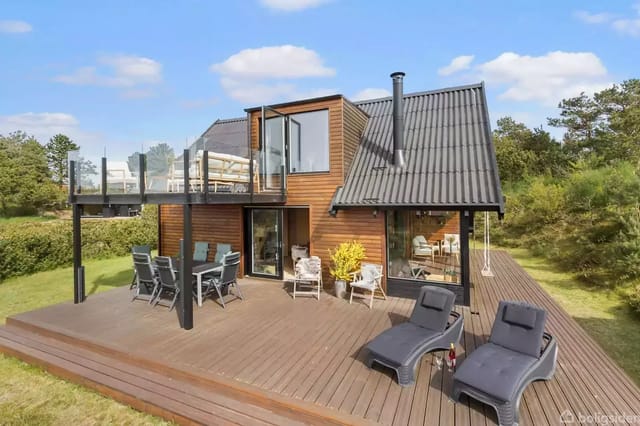 A modern holiday house with a wooden facade and black roof in a green, peaceful garden. A terrace with lounge chairs and a dining table is visible. Large windows and a roof terrace are seen.