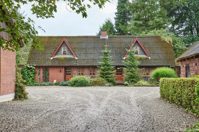 A red brick house with two pointed dormers on the roof, surrounded by trees. A gravel driveway with bushes on the side leads to the house, set against a wooded background.