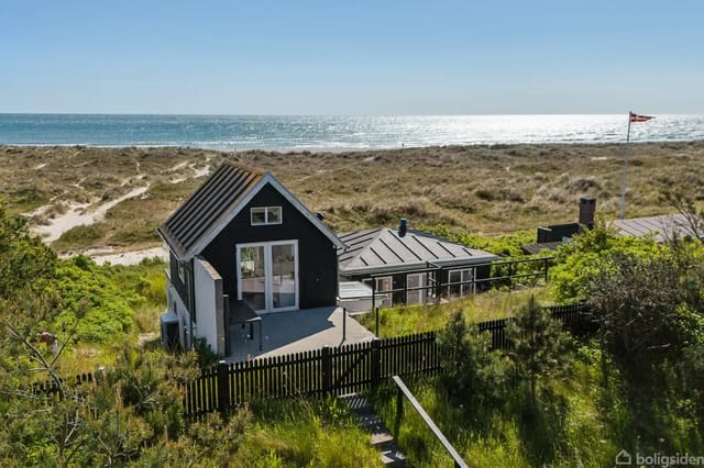 A black wooden house stands quietly on a grass-covered dune, near a sandy beach with views of the sea under a clear blue sky. A black fence surrounds the house.