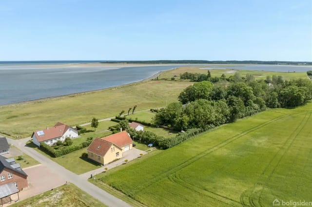 House with red roof in a quiet village near the coast, surrounded by green fields and forests, with the sea in the background under a clear blue sky.