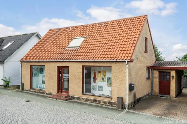 A yellow brick house with a red tiled roof and a loft window. The house has two large front windows and a red wooden door leading to a paved sidewalk.