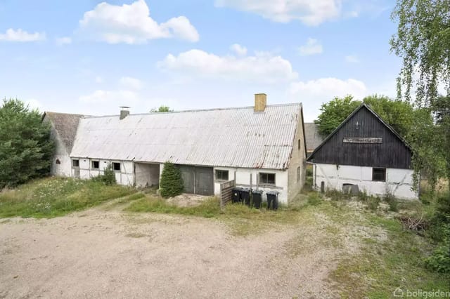 A charming old white barn with a corrugated cement roof stands next to a black wooden building, surrounded by a gravel courtyard and green trees in the background.