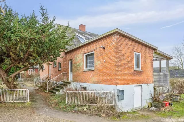 A red brick house surrounded by a gravel path and a large, dense tree. The house has a staircase at the entrance and a small shed next to it.