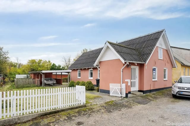 A pink house with a black roof stands quietly by a gravel road. A white picket fence encloses the garden. A carport and trees are seen in the background.