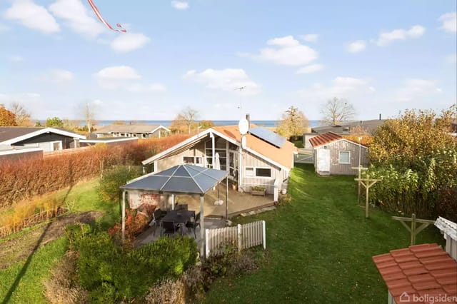 A small house stands peacefully by a green garden with hedges and trees; in the background, a blue sky and sea view.