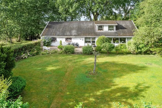 The house is set in lush garden surroundings with a lawn in front, partially surrounded by bushes and trees. It is a white single-story house with large windows and a gray roof.