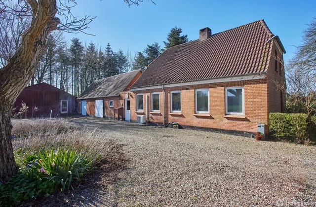 A red brick house with a sloped roof stands quietly in an open gravel area, surrounded by tall trees and bushes. There is also a large barn next to the house.