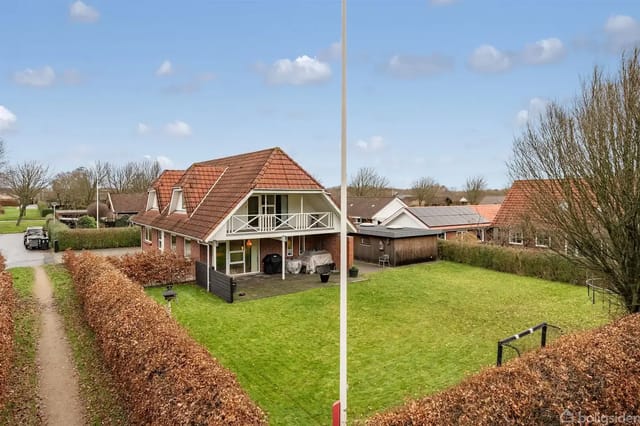Two-story house with red tile roof in a lush garden surrounded by hedges. Covered terrace in front, with neighboring houses visible.