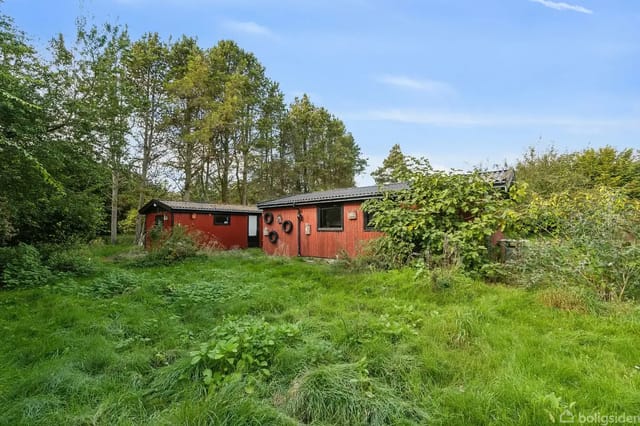 Red wooden building surrounded by tall, green vegetation in a wooded garden. Large trees form the background under a clear blue sky.