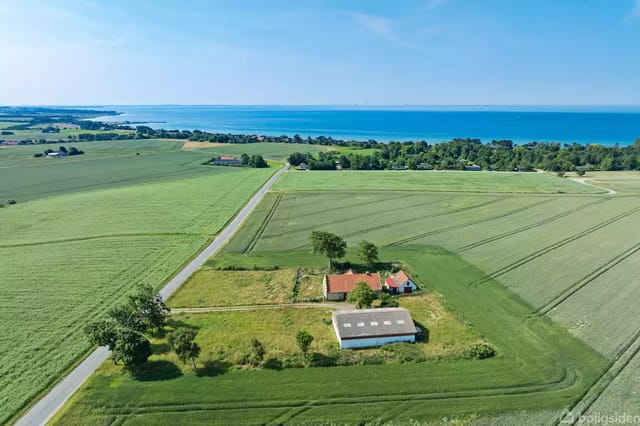 A country house with a red roof stands in the middle of a green field, surrounded by large open areas. In the background, a blue sea view under a clear sky.