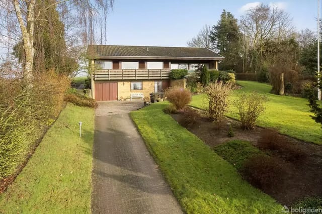 A house with a balcony is situated on an elevated plot, surrounded by a well-kept garden with shrubs and trees. A road leads up to the house, which has a garage at basement level.