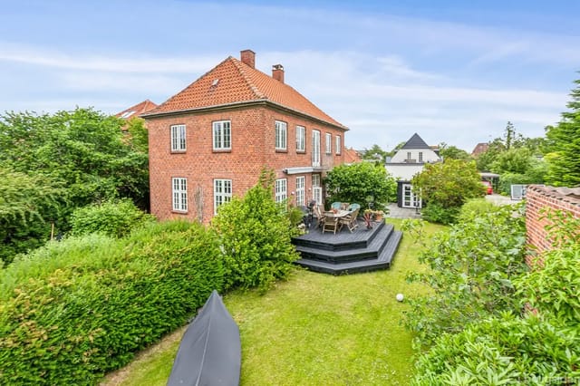 Red brick house in a green garden with terrace and garden furniture, surrounded by bushes and trees.