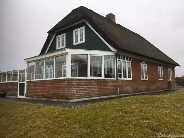 A house with a thatched roof and red brick walls stands on a lawn. A winter garden with large windows is visible. Cloudy sky in the background.