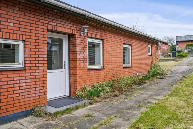 Red brick house with three windows, located by a paved path leading to a grassy area. The house has a white door and an outdoor lamp at the entrance.