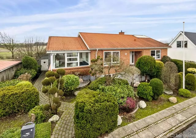 A red brick house with a red tile roof surrounded by a well-kept garden with bushes and paved paths. The house has large windows and is located in a green landscape area in a residential neighborhood.