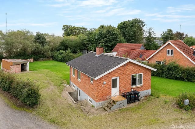 Red brick house with black roof in a green garden, surrounded by trees and other houses in a rural setting.