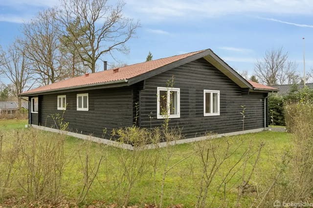 A black-painted wooden house with a red roof stands on a green lawn, surrounded by bushes and trees under a clear blue sky.