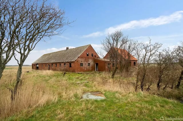 An old, red brick barn stands abandoned in an open landscape surrounded by tall grass and scattered trees under a blue sky.