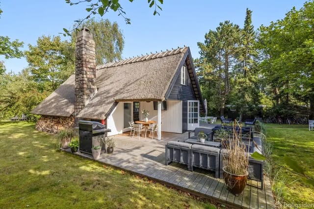 A thatched holiday home stands quietly in a lush garden with grass and trees. On the terrace, garden furniture and plants are visible as sunlight filters through the treetops.