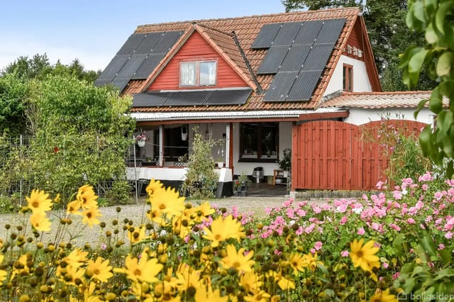 A house with solar panels on the roof, surrounded by a colorful garden with yellow and pink flowers under a blue sky.