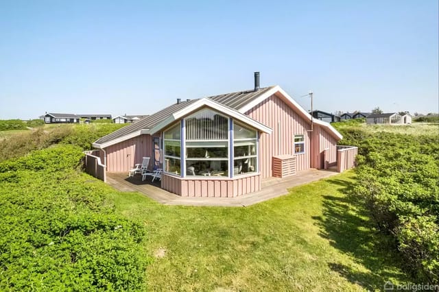 Red holiday home with large windows and terrace, surrounded by green bushes and lawn. Sunny day in a holiday home area landscape.