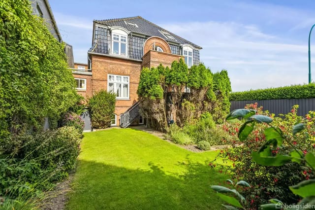 A distinctive brick house with dormer windows and a high roof, surrounded by a well-kept garden with trees and bushes in a sunny residential area.