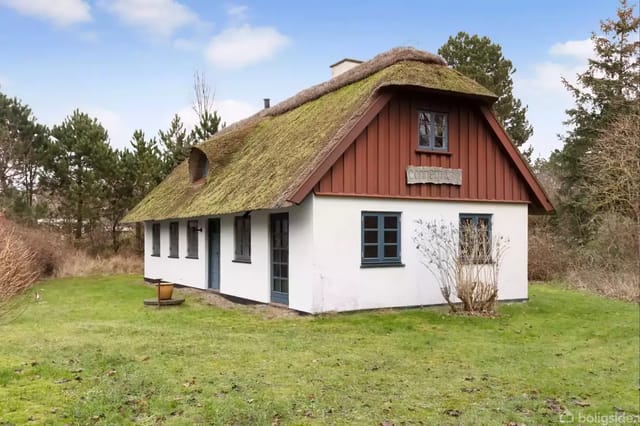 A small thatched house stands quietly on a lawn, surrounded by trees. The house has red wooden panels on the gable and white walls.