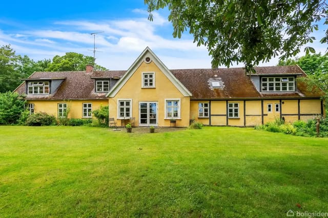 A yellow half-timbered house with brown roofs stands quietly in a lush garden. Trees surround the area under a clear blue sky.