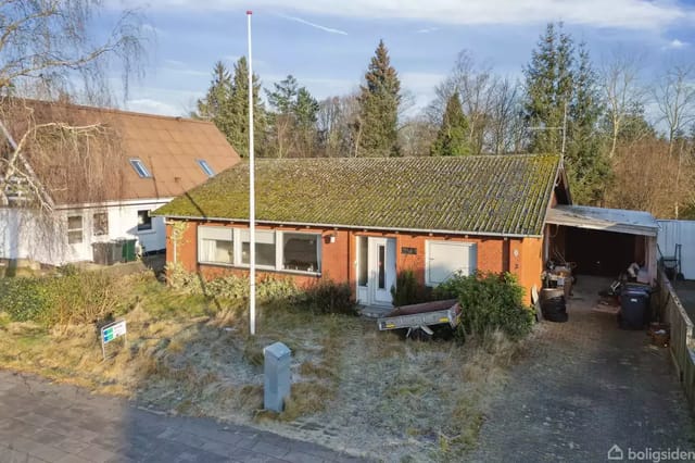 A red house with a moss-covered roof stands in a garden with a residential street in front, surrounded by tall trees. A flagpole is placed next to the house.