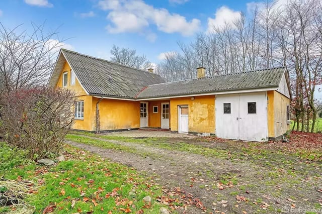 A yellow house with a gray roof stands quietly in an open, leaf-filled landscape. Forest in the background and fallen leaves on the ground create an autumn atmosphere.